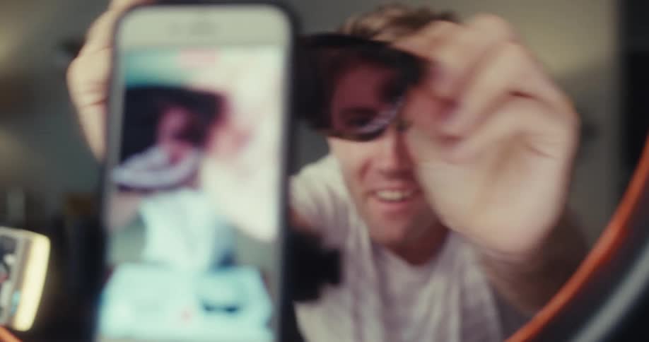 A man creates a video ad in his living room showcasing a variety of glasses while recording himself with a phone mounted on a ring light