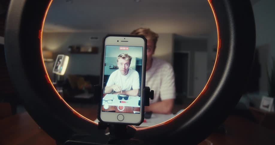 A man happily shows off his sunglasses collection filming himself with a smartphone and ring light in a cozy room