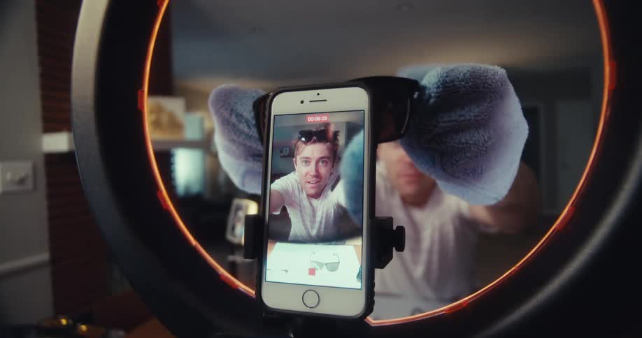 A person is recording themselves through a smartphone mounted in front of a ring light while cleaning or inspecting sunglasses