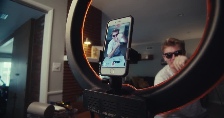 A young man enthusiastically records a video of himself dancing in front of a ring light and smartphone setup