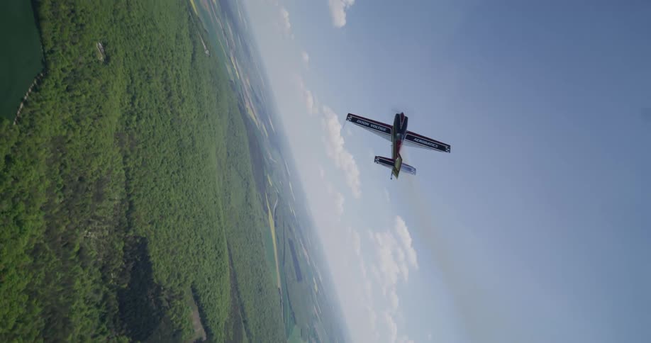Acrobatic plane flies over forest and agricultural fields leaving a trail of white smoke amidst the green landscape
