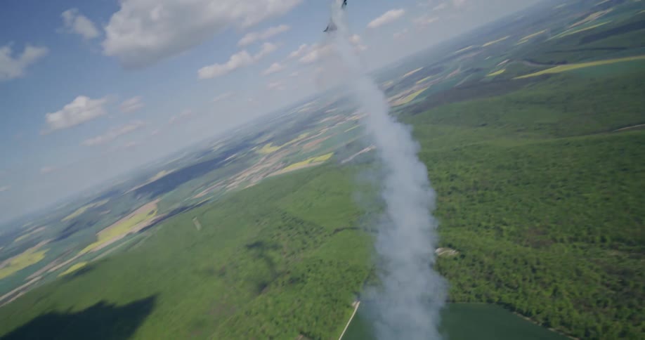An aerobatic plane soars through the sky above a patchwork of green fields and forests leaving a trail of white smoke in its wake