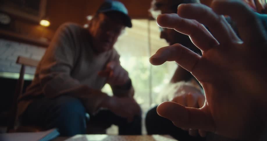 Two men are sitting indoors one wearing glasses is explaining something using hand gestures while the other wearing a hat listens attentively