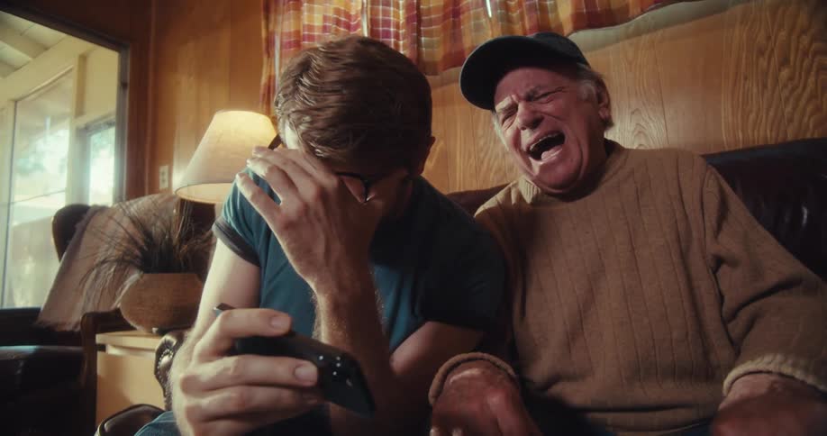 An older man wearing a veteran hat sits on a couch with a younger man in glasses both reacting intensely as they watch something on a smartphone