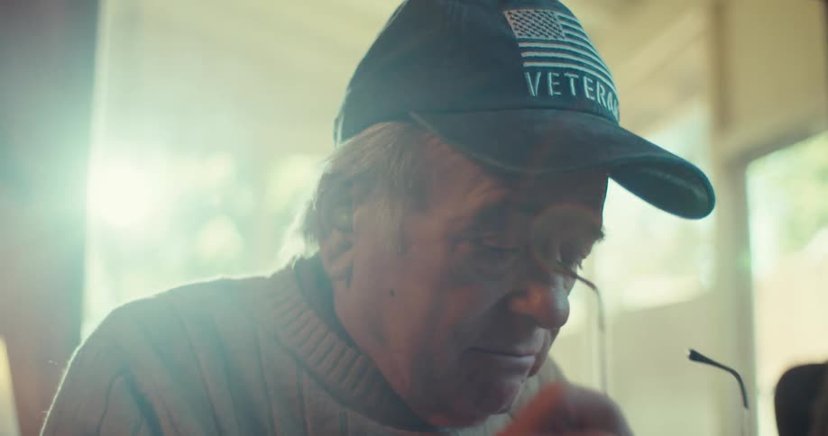 An elderly man wearing a hat takes it off speaks for a moment then puts it back on and begins writing in a notebook while sitting in his living room at midday