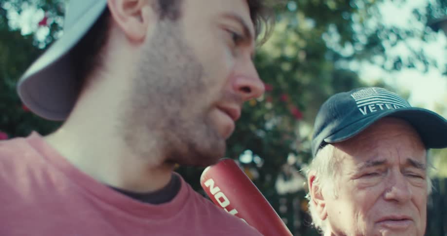 Young man with backwards cap holds baseball bat next to older veteran in outdoor conversation