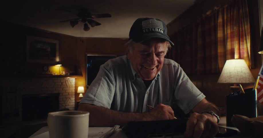 Elderly man sitting at an office desk taking off his glasses and putting them back on laughing