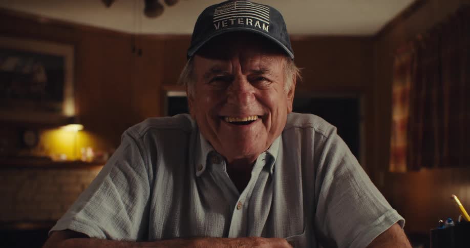 An elderly man has a big smile as he sits in his house writing with a coffee cup in front of him