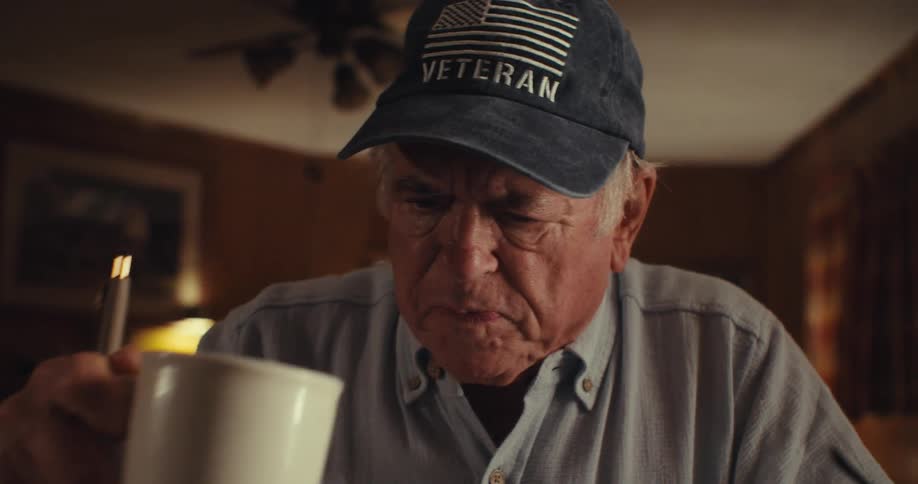 Elderly man in cap sitting drinking from a mug and holding a pen