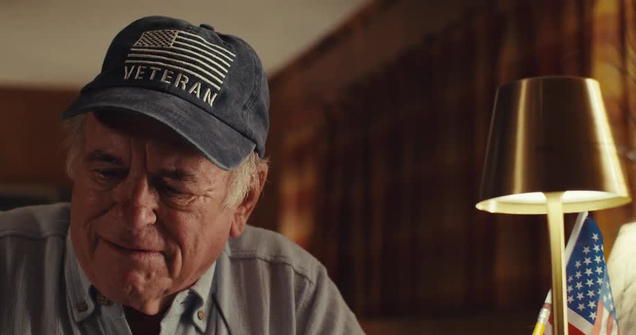 Veteran in cap and light blue shirt in a lamplit room with an american flag nearby