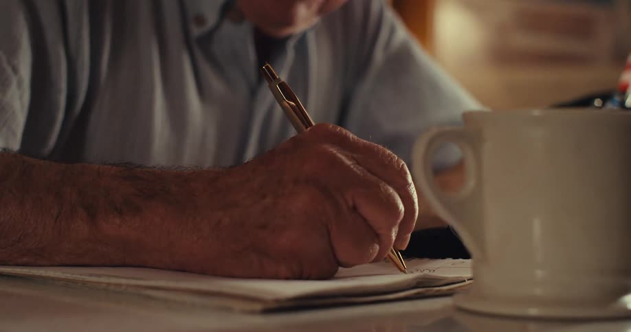 An elderly man writes in a notebook while sitting at a desk in the living room