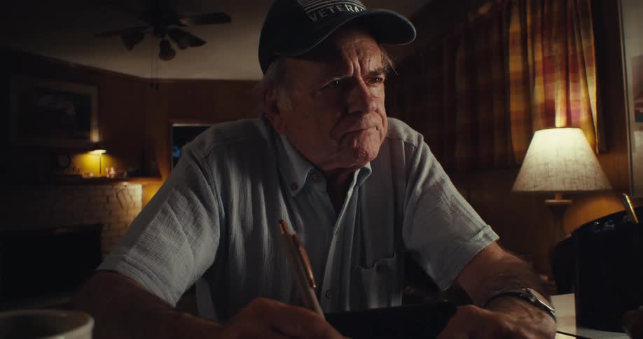 An elderly man sits behind a desk writing then suddenly takes off his glasses with a sad expression pauses briefly puts them back on and continues writing