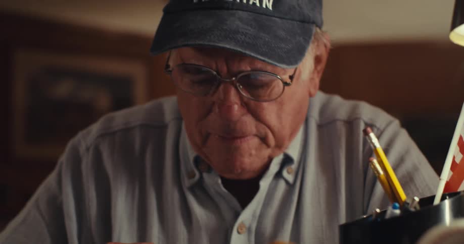 An elderly man wearing a veteran cap sits at a desk writing in a notebook with a pen looking thoughtful and perhaps troubled