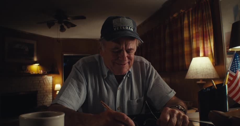 An elderly man sits behind a desk at night writing under the glow of a lamp he takes off his glasses laughs in deep thought then puts them back on and continues writing