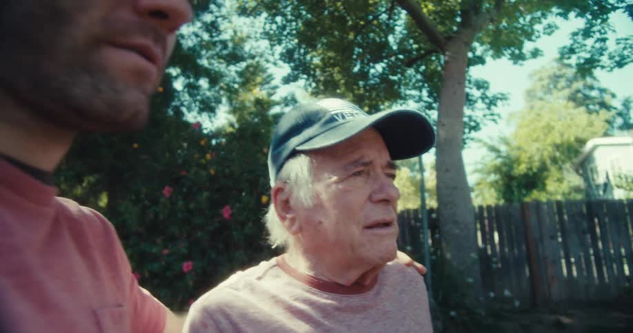 An elderly man in a veteran cap is supported by a younger man as they stand outside in a yard with trees and a fence