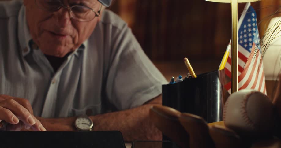 An elderly man wearing glasses sits behind a desk writing then pauses takes off his glasses and falls into deep thought for a moment before putting them back on and continuing to write