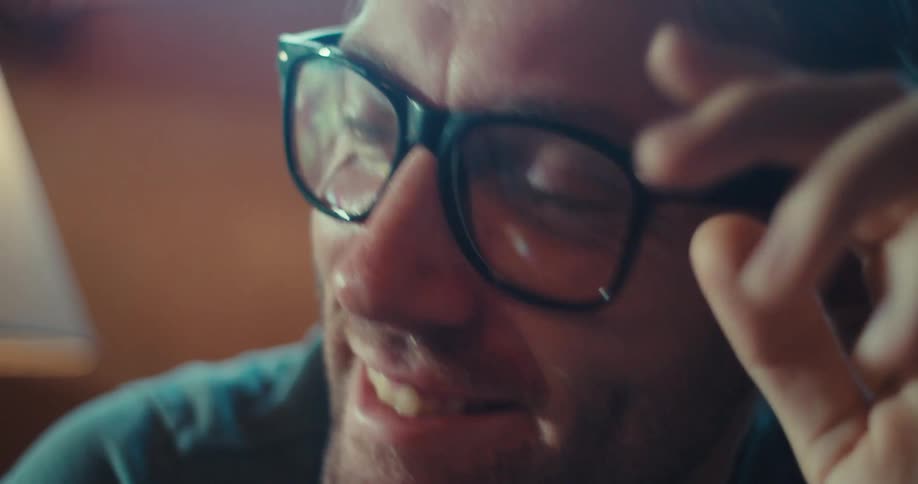 A man sitting indoors in a chair wearing glasses is deep in thought then suddenly takes off his glasses and starts laughing