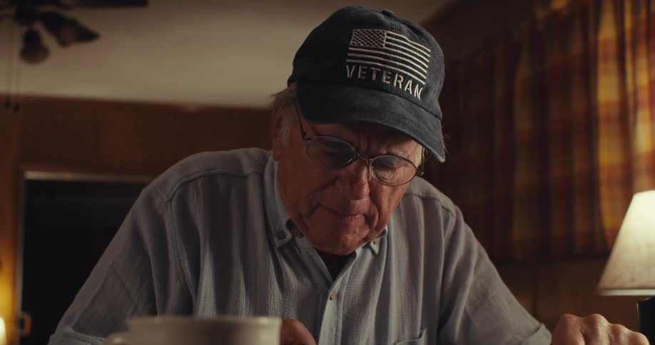 An elderly man wearing a hat and glasses sits behind a table writing