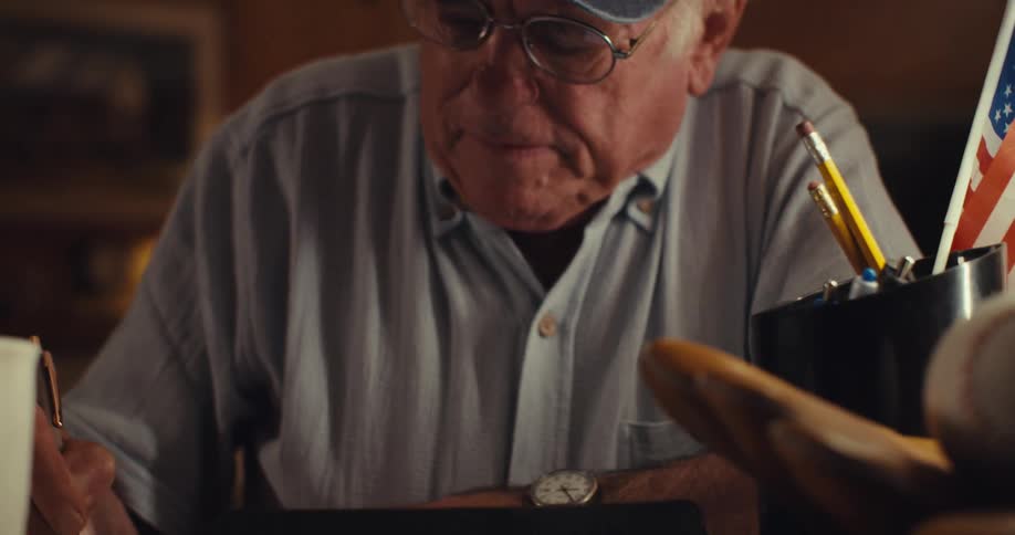 An elderly man wearing glasses and a hat sits behind a desk scrolls through a tablet then writes in a notebook