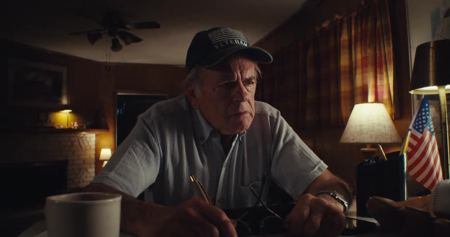 An elderly man wearing glasses sits behind a desk writing then pauses takes off his glasses and falls into deep thought for a moment before putting them back on and continuing to write