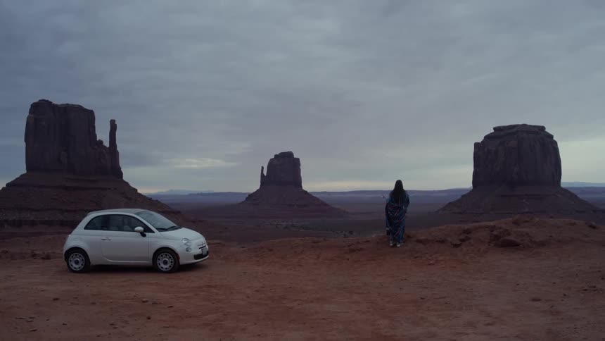 A person wrapped in a blanket walks away from their car and looks at a desert view