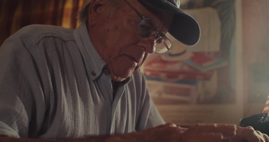 An elderly man seated behind a desk pauses his writing removes his glasses and lifts a cup of coffee to take a sip