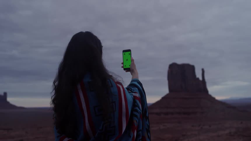 Long-haired woman holding her smart phone out in front of her pointed towards some large rock formations on a very cloudy day