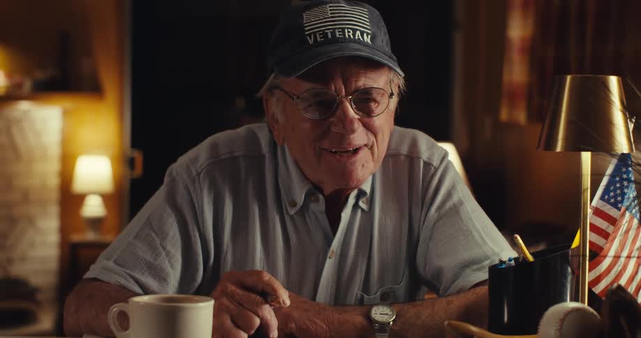 Smiling veteran wears a cap and lightcolored shirt as he talks in a cozy room with an american flag on the table