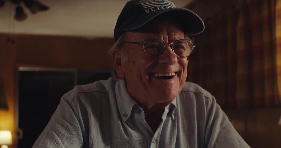 An elderly man sitting behind a desk wearing a hat and glasses suddenly starts laughing after a moment of deep thought