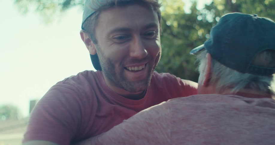 A young man talking and tossing a baseball up and down then joyfully hugging an elderly man beside him on their lawn at midday