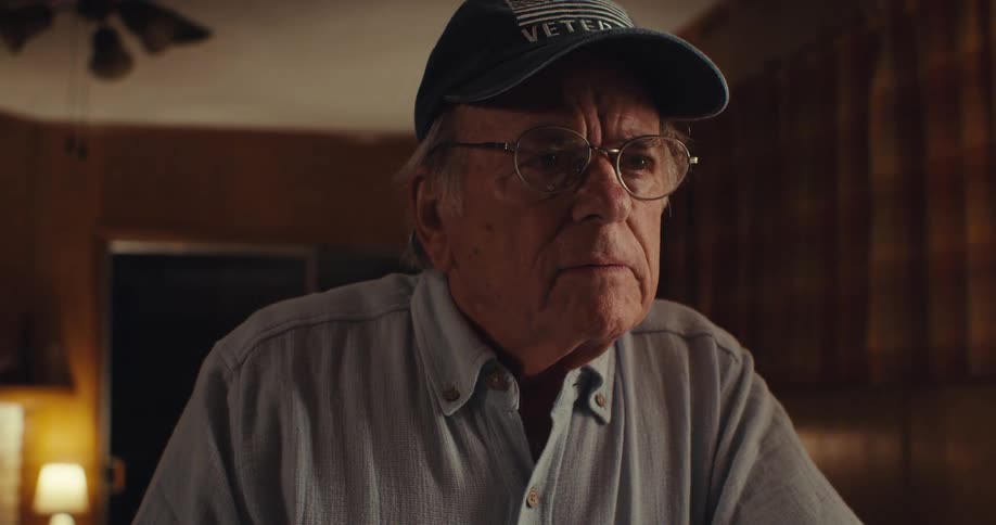 An elderly man wearing glasses and a veteran cap appears deep in thought in a warmly lit home interior