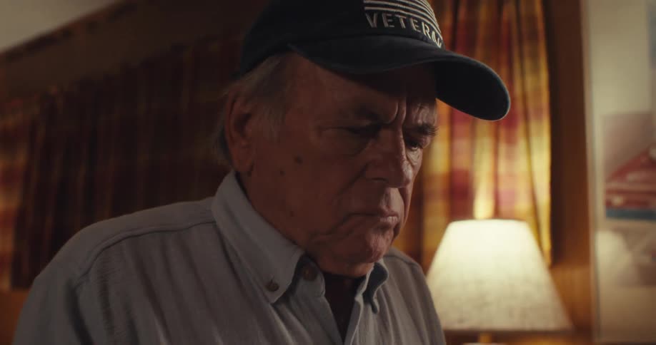 Man in veterans cap looks with serious expression while sitting in a room