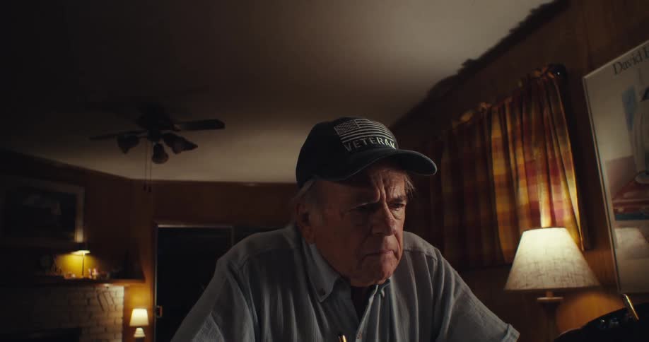 An elderly man sitting behind a desk removes his glasses while in deep thought for a moment then puts them back on and continues writing