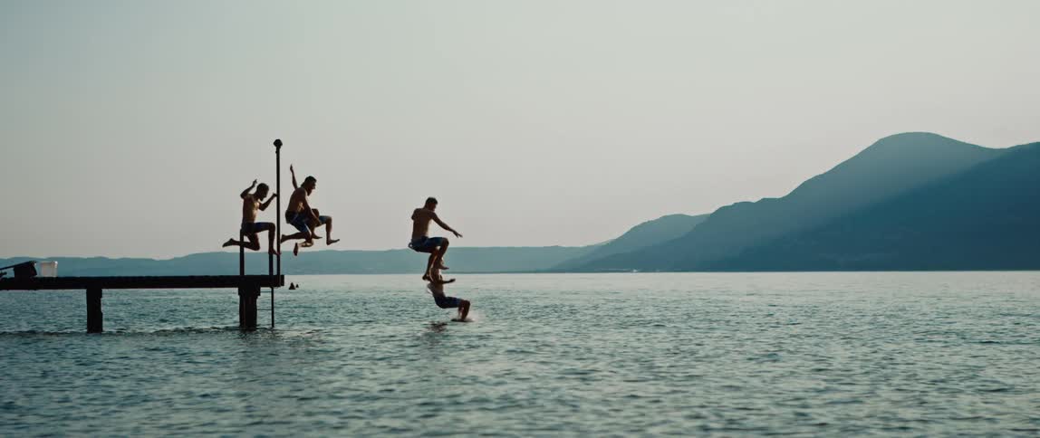 Group of friends running on wood dock and jumping into lake at dusk