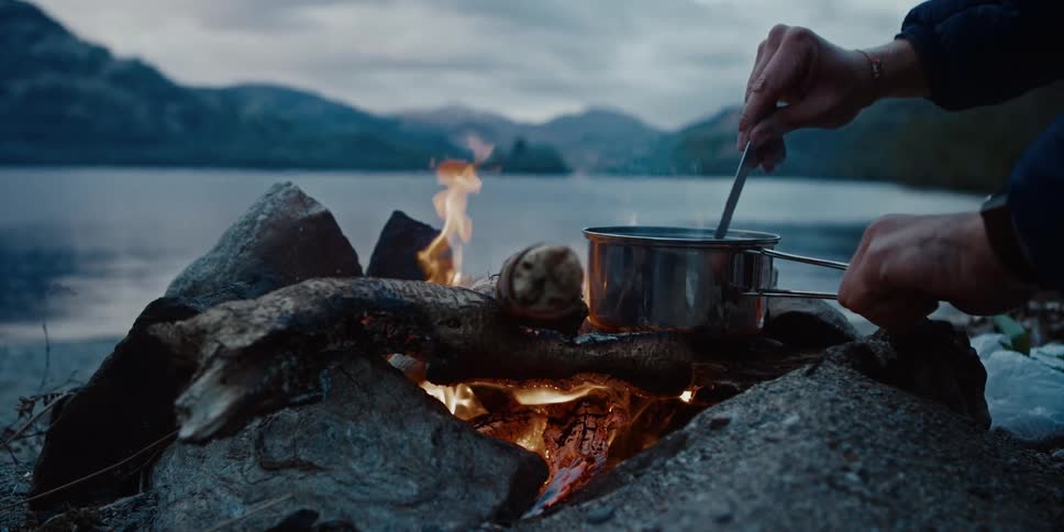 Person cooks in a pot over a fire on the edge of a lake surrounded by mountains at dusk