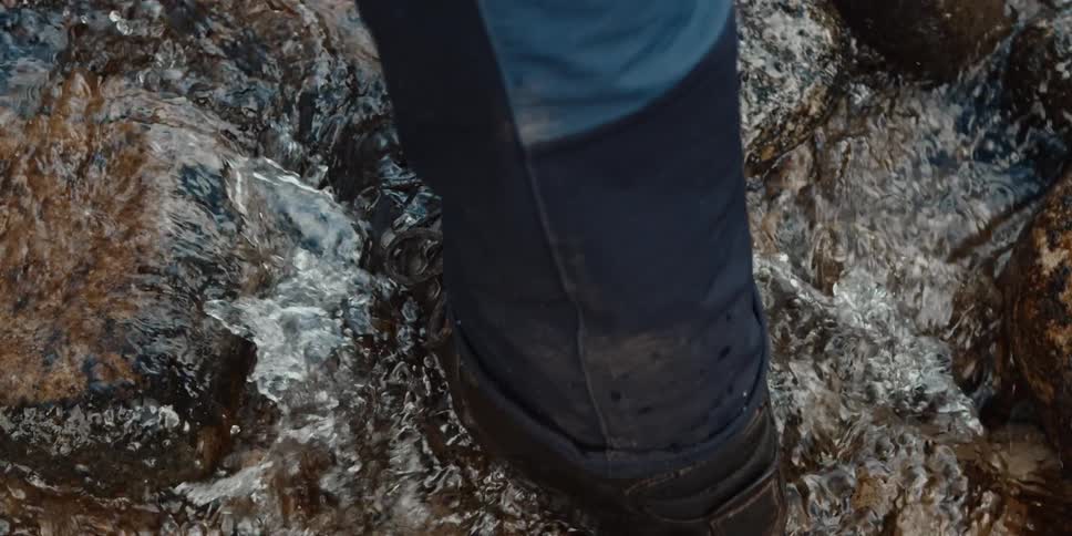A hikers boot splashes through a shallow rocky stream in slow motion
