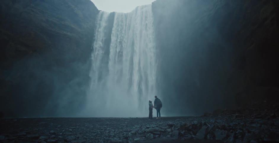 Adult and child stand looking up at a waterfall near a misty cliff during the daytime