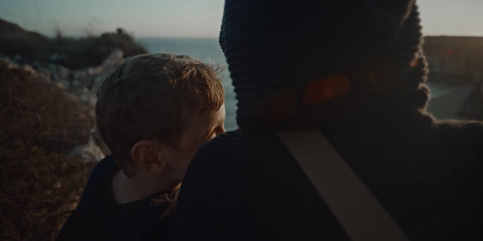 Child eating looks on intently next to a hooded person as they sit near the sea during sunrise