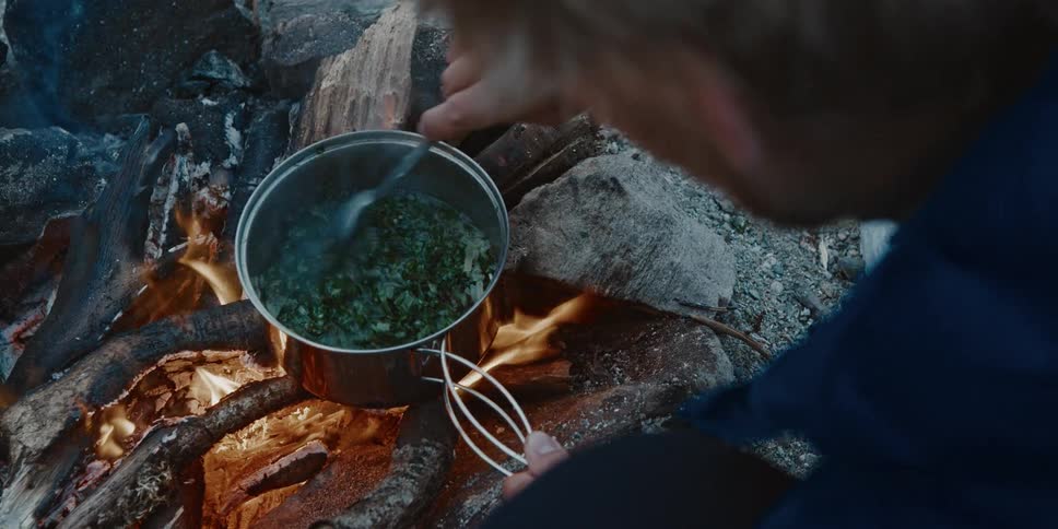 Man cooks herbs in a pot over a wood fire at an outdoor campsite