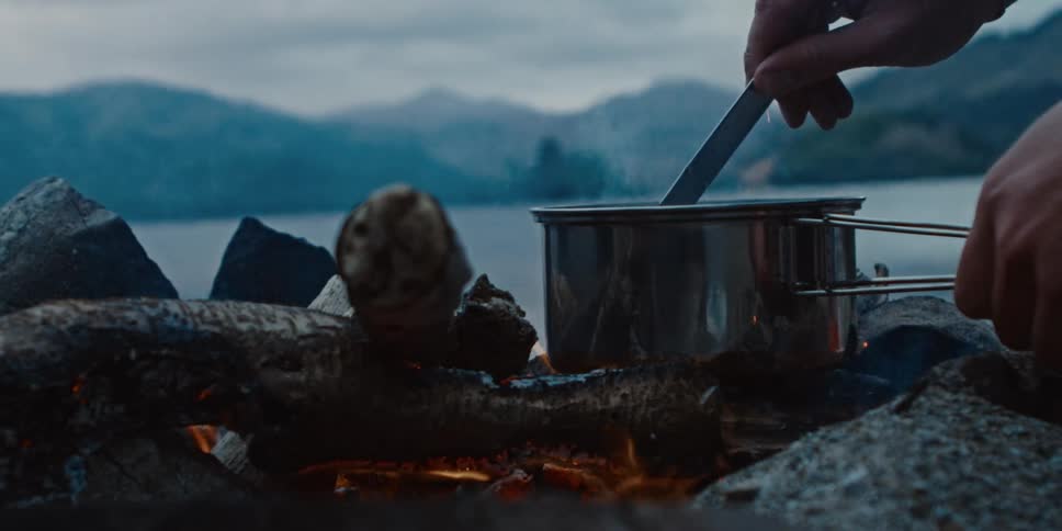 Stirring spoon in a pot over a campfire while camping overlooking lake and mountains