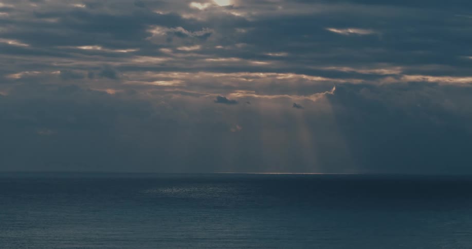 Beams of light pierce through dense clouds and gently illuminate the sea surface during dusk