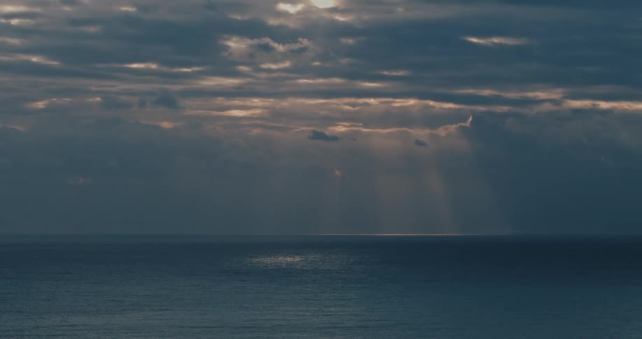 A view of the surface of an ocean with the suns rays coming from the sky that is full of clouds