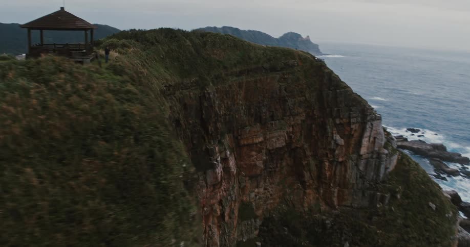 Person stands near a building built on a seaside cliff while waves crash against the rocks