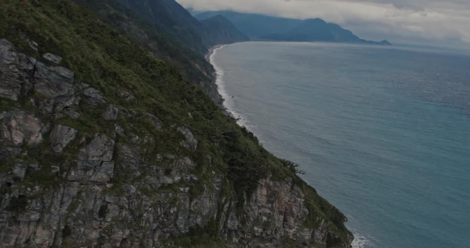 Hillside covered in dense forest follows a strip of rocky coastline under a cloudy sky and mountains in the background