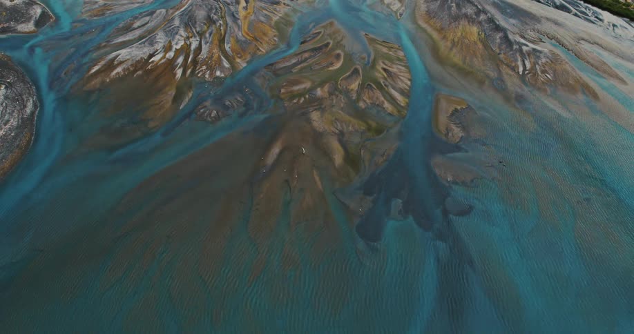 An aerial view shows a braided river system with turquoise water flowing through sediment deposits of various colors