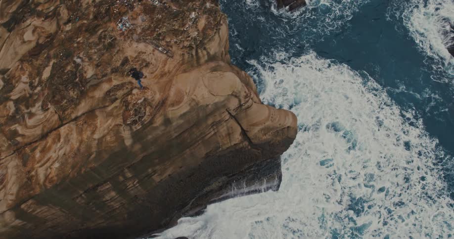 Person with a backpack stands on a seaside cliff as the waves crashing against its base foam and churn