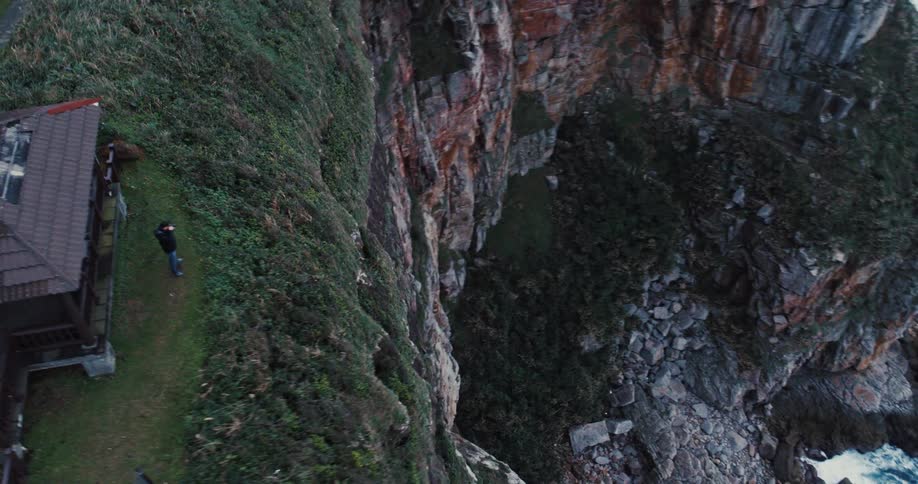 Aerial shot of a person standing in front of a house on the edge of a cliff on a mountain by the sea