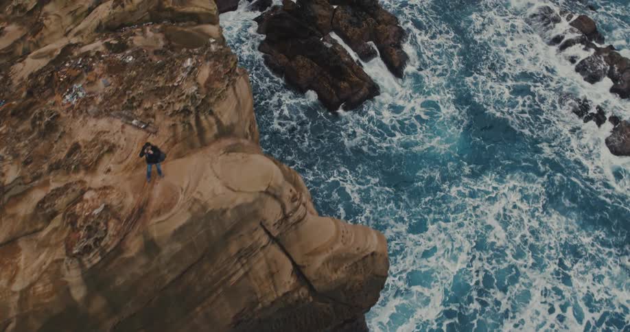 Man standing at the edge of a promontory as waves roll in and splash against rocks below
