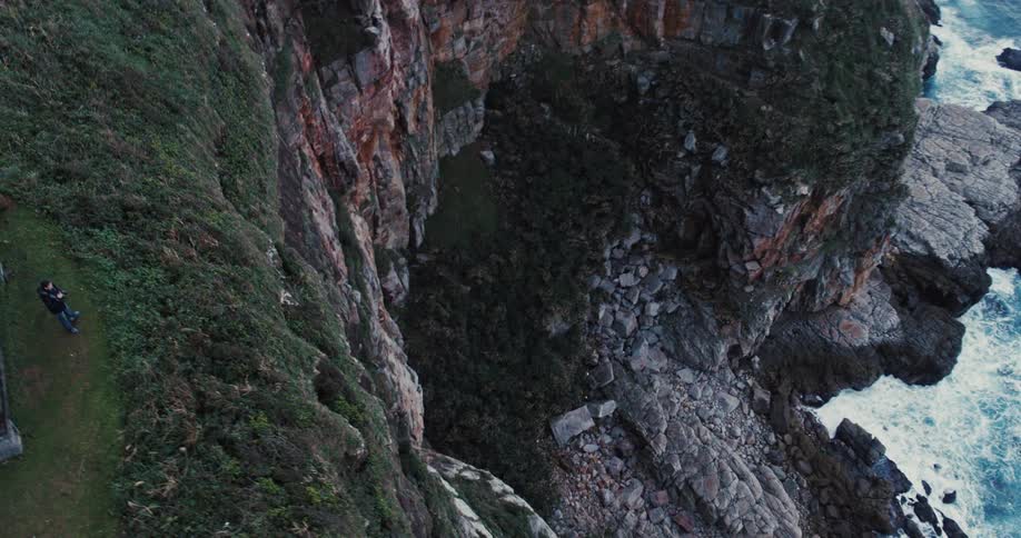 Aerial shot of a person standing next to a house on a mountain peak at the edge of a cliff and the sea