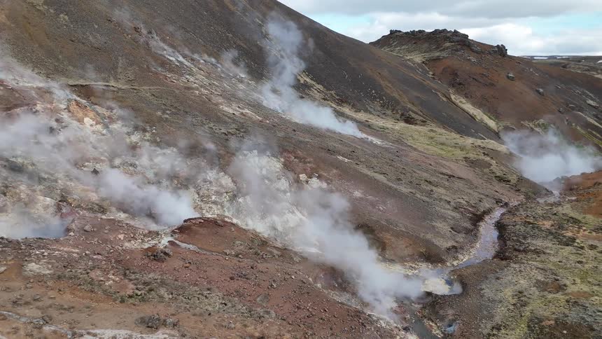 Vapors rise from fissures in the ground on a volcanic slope with areas of earthy rocks and sparse vegetation